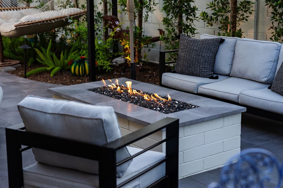 Modern outdoor patio with fire pit, gray seating, and lush greenery in a cozy setting.