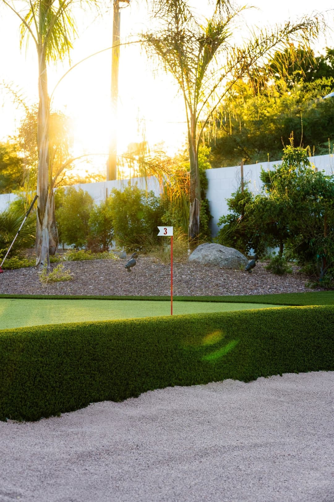 Golf course with sand trap and flag number 3 during sunset, surrounded by palm trees.