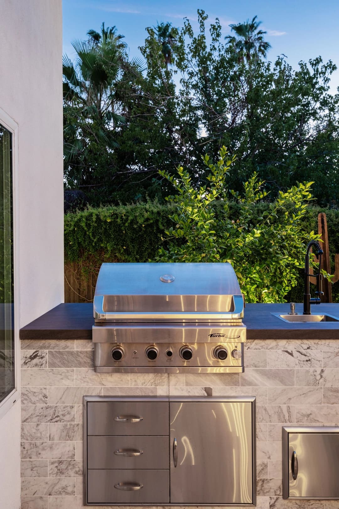Stainless steel outdoor grill with marble countertop and lush garden background at dusk.