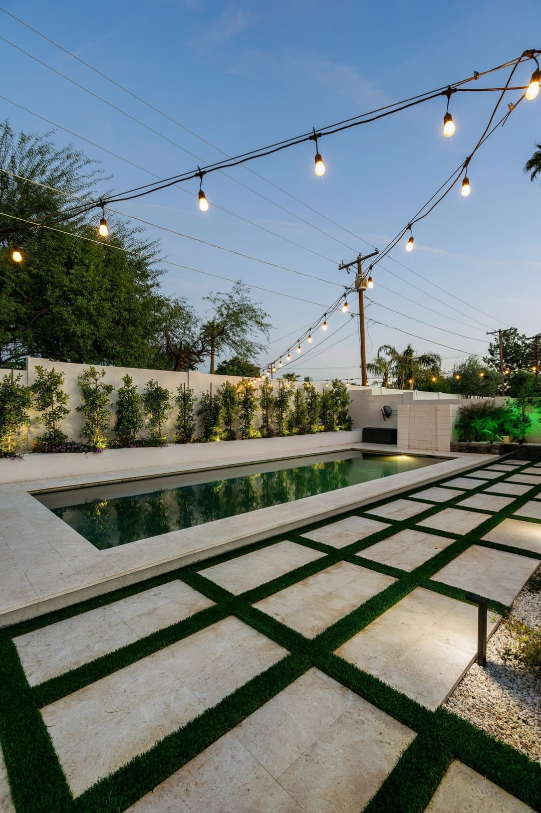 Modern backyard pool with stylish landscaping, illuminated by string lights at dusk.