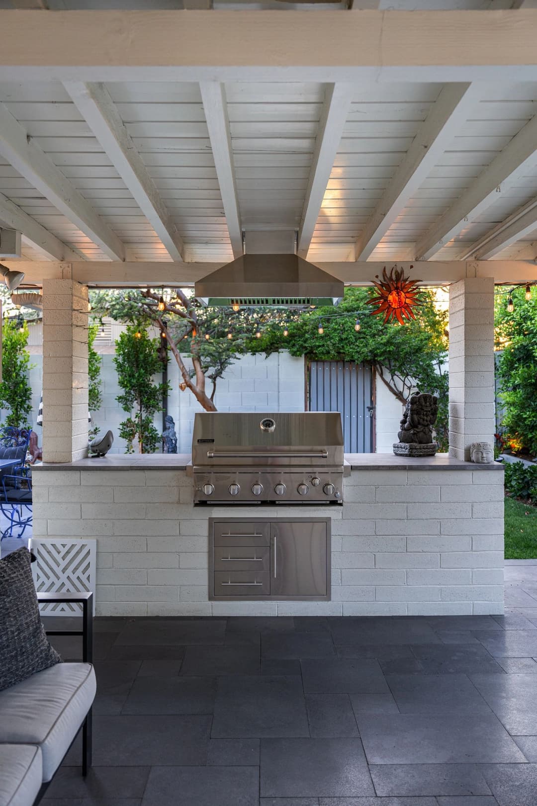Outdoor kitchen with a stainless steel grill under a covered patio surrounded by greenery.
