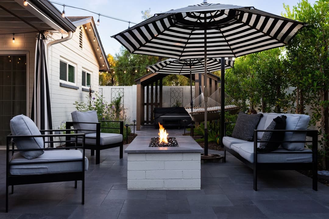 Cozy outdoor patio with fire pit, modern seating, striped umbrella, and lush greenery.
