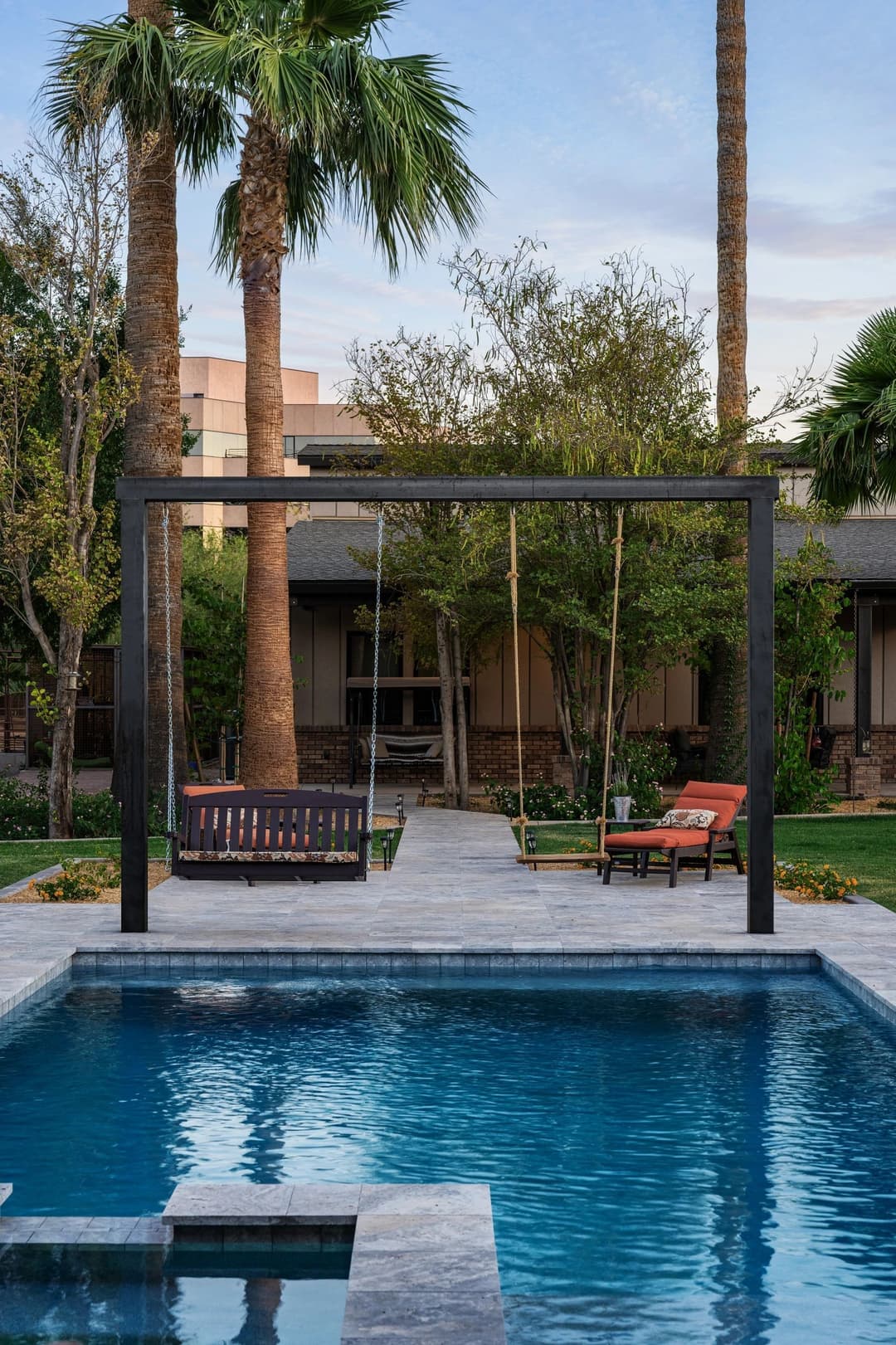 Elegant backyard oasis with a swimming pool, swings, and palm trees under a clear sky.