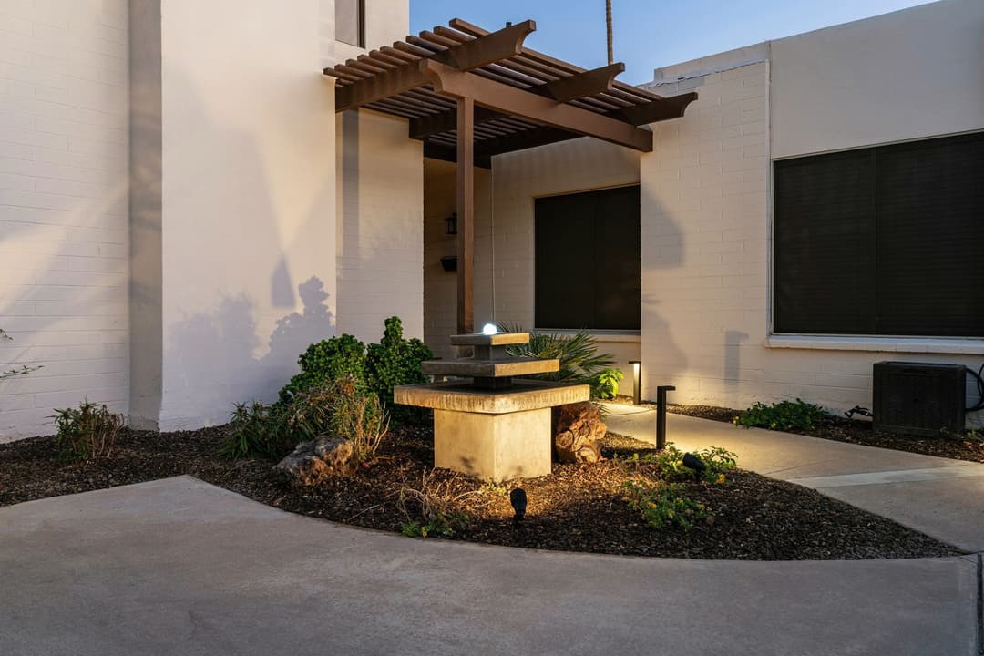 Modern outdoor entrance with stone fountain, lush greenery, and warm lighting at dusk.