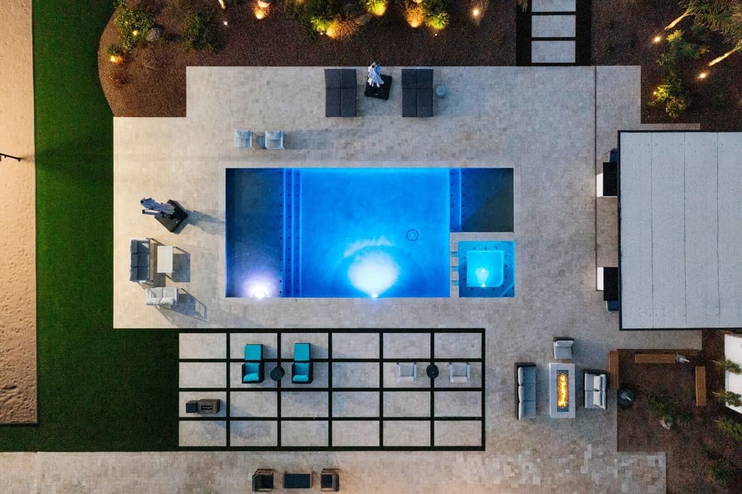 Aerial view of a modern backyard pool with spa, lounge chairs, and fire pit at dusk.