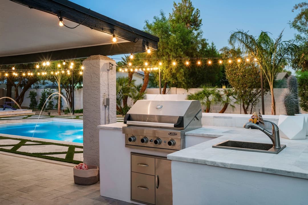 Modern outdoor kitchen with stainless steel grill, elegant pool, and string lights in a serene backyard.