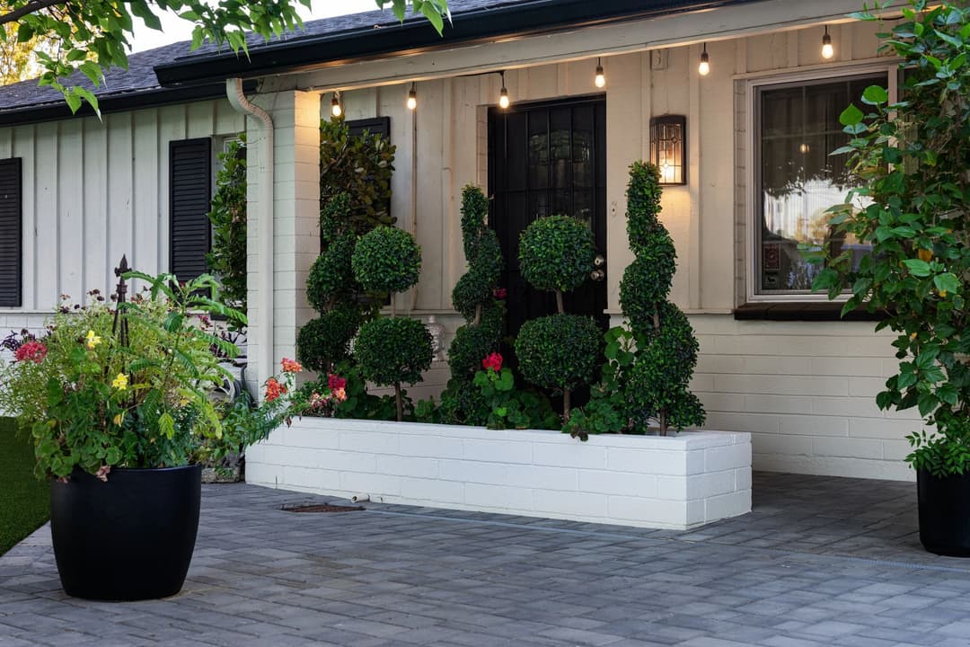 Charming front porch with topiary shrubs, flowers, and warm string lights in a cozy home setting.