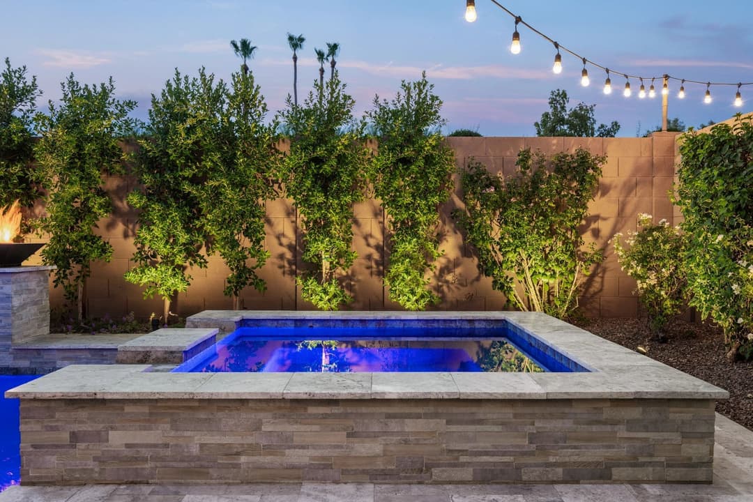 Elegant backyard spa with blue water, surrounded by illuminated greenery and evening sky.