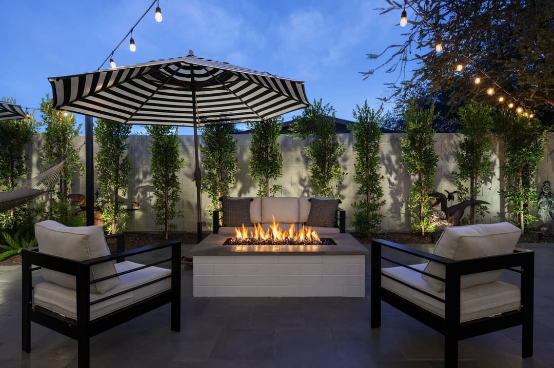 Cozy backyard fire pit with seating under a striped umbrella and string lights at dusk.