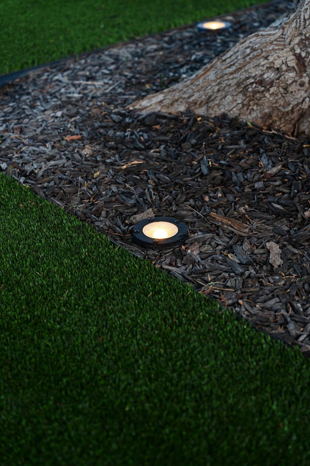 Outdoor landscape lighting near a tree on mulch and artificial grass. Safe and stylish illumination.